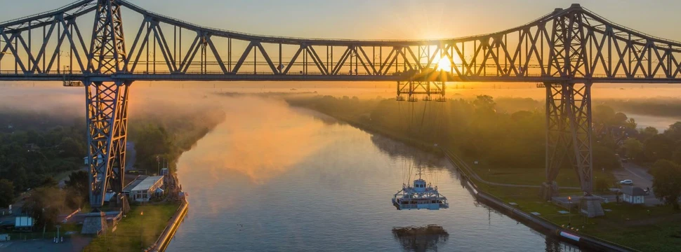 Eine metallerne Brücke in Rensburg über dem Nord-Ostsee-Kanal mit der Schwebefähre an der Unterseite und dem Sonnenuntergang im Hintergrund