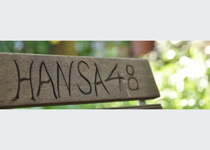 Bank mit der Inschrift "HANSA48" in einem begrünten Hinterhof. Holzbank mit eingeritztem Schriftzug "HANSA48" im sonnigen Garten.