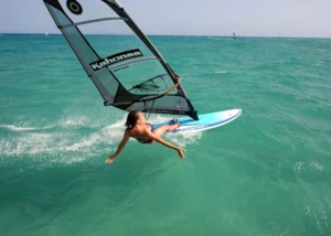 Dynamische Windsurf-Action auf dem offenen Meer. Windsurferin hängt seitlich am Segel und gleitet rasant über türkisblaues Meer.