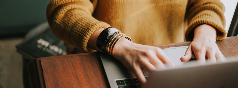 Eine Person, die in einem gelben Pullover vor einem Tisch sitzt und an einem Laptop arbeitet