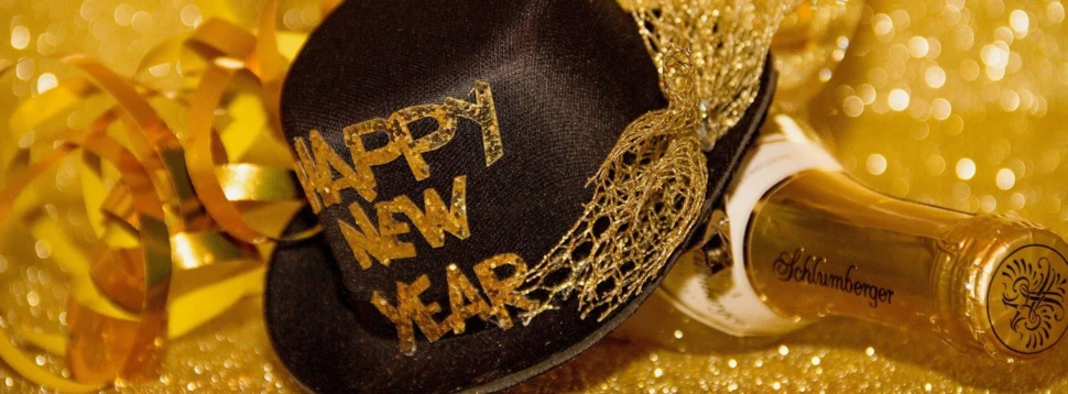 Partyhut mit Happy New Year Aufschrift an einer Sektflasche vor goldenem Hintergrund.