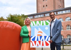 Eine Frau und ein Mann, die vor dem Kieler Rathaus stehen und das Logo der Kieler Woche 2026 präsentieren
