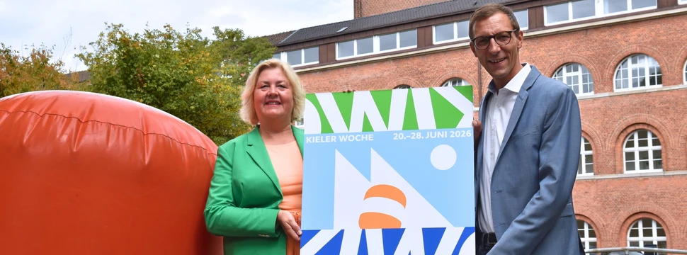 Eine Frau und ein Mann, die vor dem Kieler Rathaus stehen und das Logo der Kieler Woche 2026 präsentieren