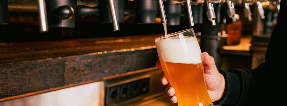 Hand hält frisch gezapftes Bier mit Schaum vor glänzenden Zapfhähnen einer modernen Bar.