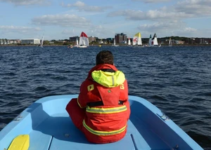 Ein Mensch in einer orangenen Warnweste in einem blauen Boot auf dem Wasser der Kieler Förde mit Segelbooten im Hintergrund