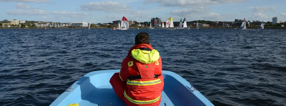 Ein Mensch in einer orangenen Warnweste in einem blauen Boot auf dem Wasser der Kieler Förde mit Segelbooten im Hintergrund