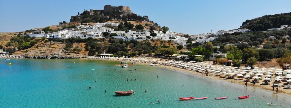 Strand mit Berg im Hintergrund. Im Wasser sieht man Menschen und Surfboards