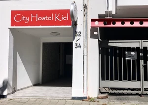Eingang des City Hostel Kiel bei sonnigem Wetter. Eingang des City Hostel Kiel mit rotem Schild und einer Toranlage daneben bei Sonnenschein.