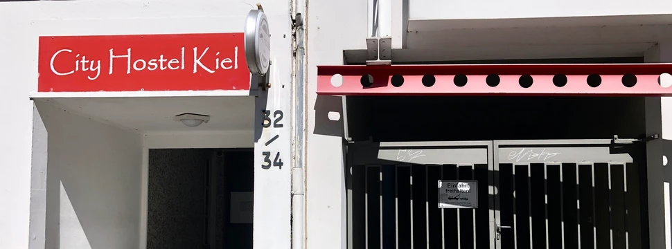 Eingang des City Hostel Kiel mit rotem Schild und einer Toranlage daneben bei Sonnenschein.