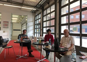 Gesprächsrunde in der lichtdurchfluteten Stadtgalerie. Drei Personen sitzen an runden Tischen in einer hellen Stadtgalerie mit großen Fenstern und modernem Design.