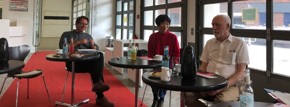 Drei Personen sitzen an runden Tischen in einer hellen Stadtgalerie mit großen Fenstern und modernem Design.
