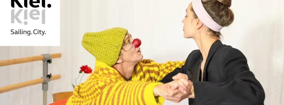 Zwei Frauen, davon eine in einem Clownskostüm, tanzen gemeinsam in einem Raum mit weißen Wänden