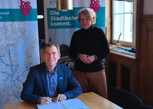 Zwei Menschen vor Bannern, die gerade gemeinsam einen Vorvertrag unterschrieben haben für eine kommende Straßenbahnstrecke