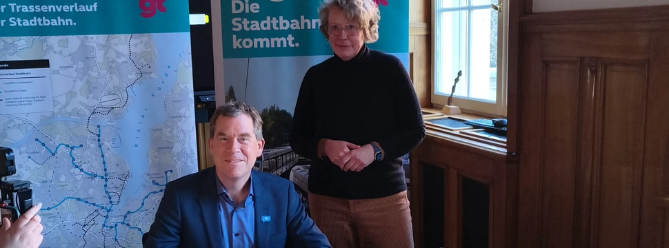 Zwei Menschen vor Bannern, die gerade gemeinsam einen Vorvertrag unterschrieben haben für eine kommende Straßenbahnstrecke