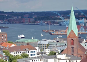 Panoramablick auf Kiel mit Nikolaikirche, Förde, Schiffen und Windrädern am Horizont bei bewölktem Himmel.