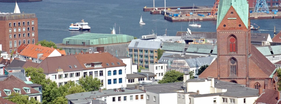 Panoramablick auf Kiel mit Nikolaikirche, Förde, Schiffen und Windrädern am Horizont bei bewölktem Himmel.