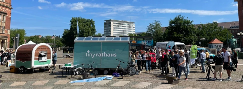 Ein türkiser Anhänger mit "tiny rathaus" Aufschrift und Solarpanels auf einem Stadtplatz, umgeben von Bürgern.