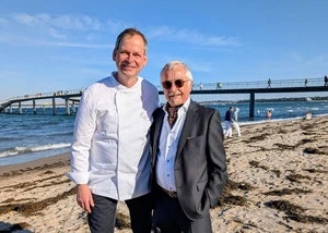Christoph Rüffer und Klaus-Peter Wilhöft bei der Eröffnung der 39. Ausgabe des Schleswig-Holsteinischen Genussfestivals am Strand von Travemünde, Zwei Männer unterschiedlichen Alters an einem Strand mit Wasser und einer Seebrücke im Hintergrund