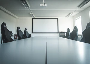 Eine Leinwand hinter einem langen weißen Tisch, an dem mehrere Schreibtischstühle stehen