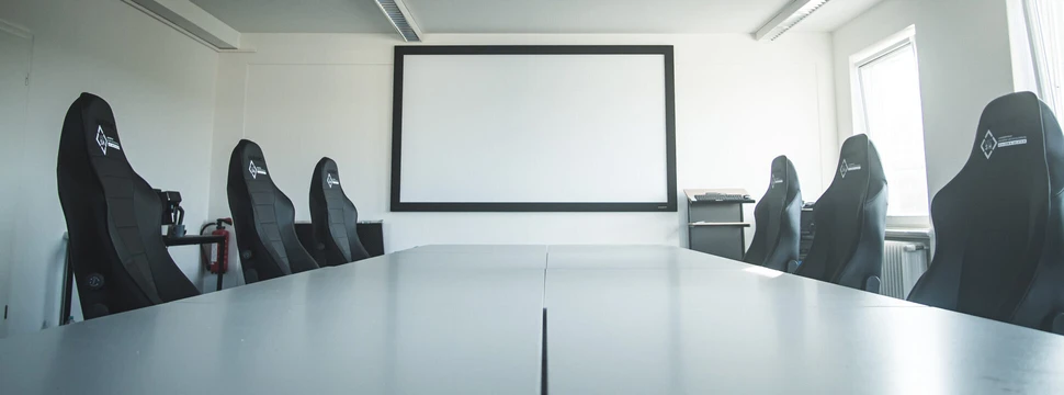 Eine Leinwand hinter einem langen weißen Tisch, an dem mehrere Schreibtischstühle stehen