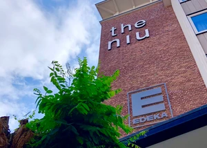 Ein modernes Wohngebäude "the niu" mit charakteristischer Backsteinarchitektur und urbaner Begrünung. Modernes Backsteingebäude mit "the niu" Schriftzug und grünem Baum im Vordergrund bei blauem Himmel.