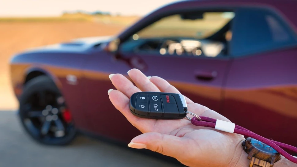 Hand hält schwarzen Autoschlüssel mit rotem SUV im Hintergrund bei warmem Abendlicht