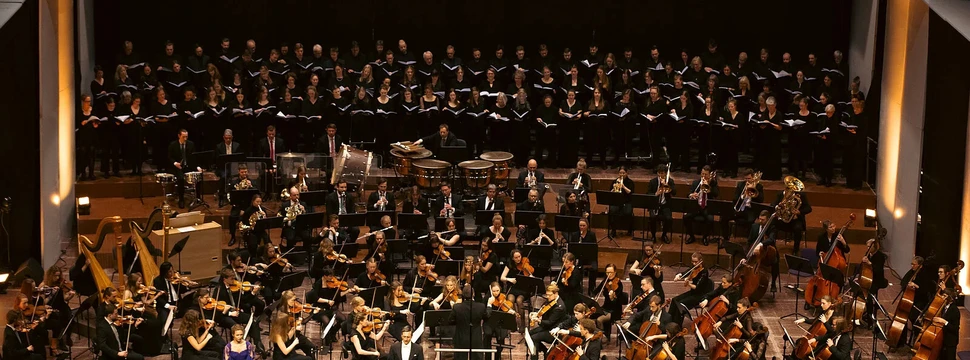 Ein Orchester mit einem Chor auf einer großen Bühne, die gerade einen Auftritt haben