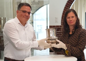 Ali Arseven von der Fielmann Group und Dr. Sonja Kinzler als Leiterin des Kieler Stadt- und Schifffahrtsmuseums bei der Übergabe des Pokals Ein Mann in einem Hemd und eine Frau ebenfalls in einem Hemd gekleidet mit einem Pokal aus Silber in den Händen in einem Kieler Museum