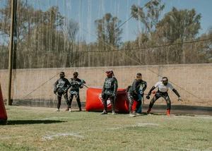 Eine Paintballmannschaft beim Aufwärmen Mehrere Menschen in Schutzkleidung und Paintballequipment vor einer Mauer und einem orangenem Luftkissen