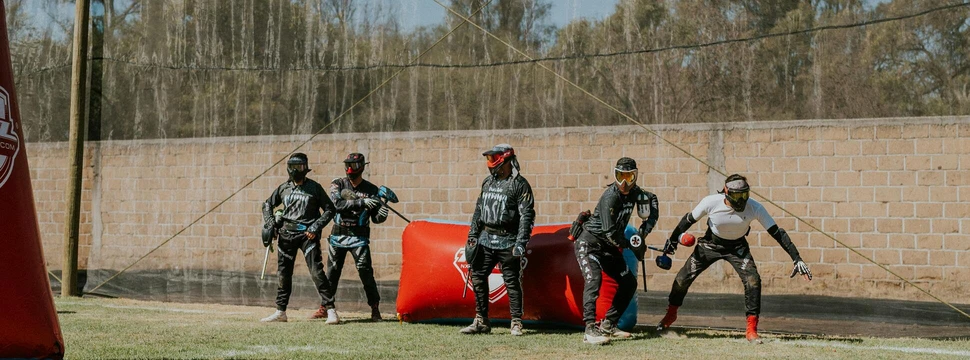 Mehrere Menschen in Schutzkleidung und Paintballequipment vor einer Mauer und einem orangenem Luftkissen