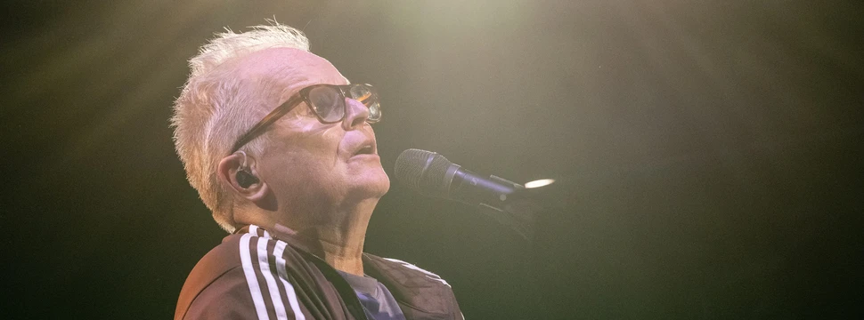 Ein Mann mit grauen Haaren vor einem Mikrofon auf einer Bühne mit einem Scheinwerfer im Hintergrund