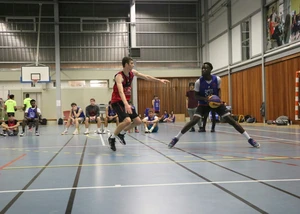 Zwei junge Männer spielen in einer Sporthalle Handball. Im Hintergrund sieht man weitere Jugendliche auf einer Bank sitze.