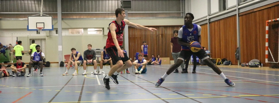 Zwei junge Männer spielen in einer Sporthalle Handball. Im Hintergrund sieht man weitere Jugendliche auf einer Bank sitze.