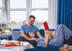 Frau liegt mit dem Kopf auf dem Schoß von einem Mann. Sie hat ein Buch in der Hand und lacht ihren Partner an