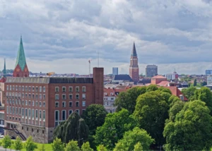 Kieler Innenstadt mit Rathausturm und Nikolaikirche im Sommerlicht. Skyline von Kiel mit Rathausturm, Nikolaikirche und grünen Baumkronen unter leicht bewölktem Himmel.