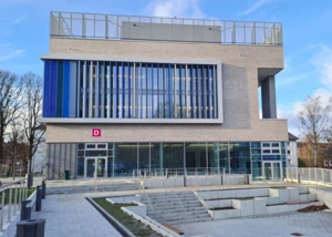 Urbaner Neubau mit Glasfassade und Eingangsbereich D. Moderner Gebäudeeingang mit Glasfassade und blauen Lamellen am Abschnitt D.