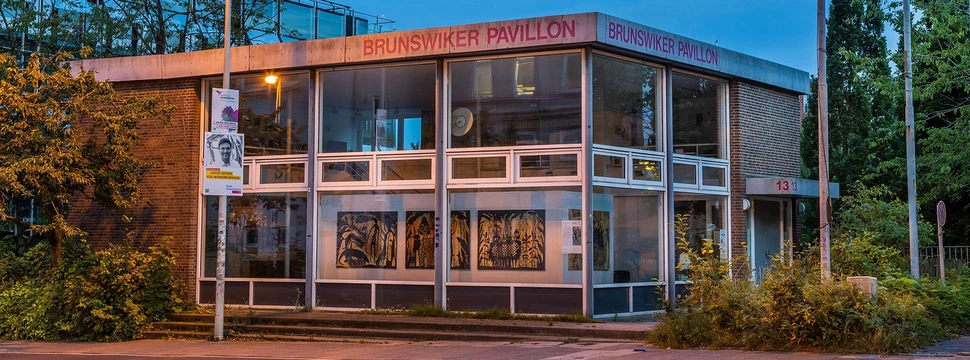 Ein Gebäude mit einer breiten Fensterfront mit Bäumen im Hintergrund, an dessen Fassade "Brunswiker Pavillon" steht