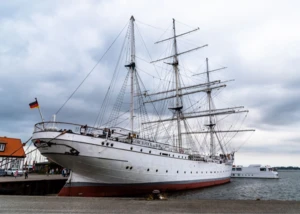 eißes Segelschiff Gorch Fock mit drei Masten und deutscher Flagge liegt am Hafenkai bei bewölktem Himmel