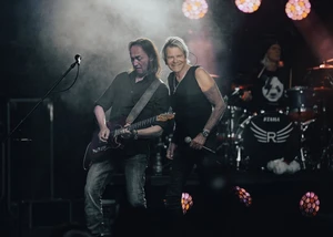 Zwei Männer sind auf einer Bühne. Einer spielt auf einer Gitarre der Andere hält ein Mikrofon in der Hand. Im Hintergrund befindet sich ein Drummer.