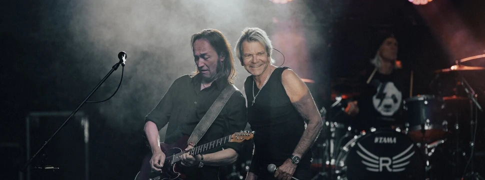 Zwei Männer sind auf einer Bühne. Einer spielt auf einer Gitarre der Andere hält ein Mikrofon in der Hand. Im Hintergrund befindet sich ein Drummer.