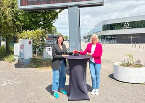 Zwei Frauen vor einem mit schwarzer Decke, auf dem ein roter Buzzer steht, mit dem ein neues kostenloses Kulturangebot hingewiesen wird