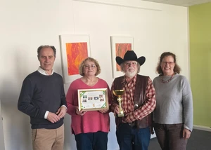 Vier Menschen vor einer weißen Wand, die gerade eine Urkunde und einen Pokal übergeben bzw. erhalten haben