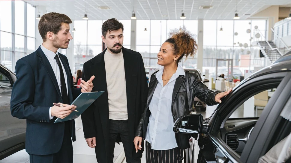 Autoverkäufer berät Paar im modernen Showroom neben schwarzem Auto mit Klemmbrett in der Hand.