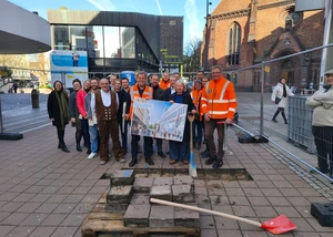 Mehrere Menschen, einige davon mit einer orangenen Warnweste, vor einer Baustelle in der Kieler Innenstadt, deren geplante Gestaltung auf einem Plakat zu sehen ist, das von einer Person in der Mitte des Bildes gehalten wird