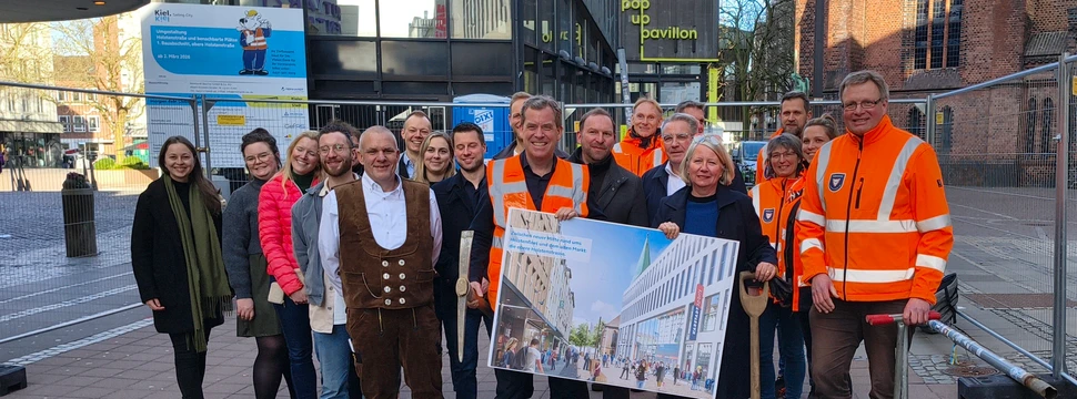 Mehrere Menschen, einige davon mit einer orangenen Warnweste, vor einer Baustelle in der Kieler Innenstadt, deren geplante Gestaltung auf einem Plakat zu sehen ist, das von einer Person in der Mitte des Bildes gehalten wird