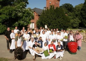 Eine Gruppe von Menschen in Schürzen mit regionalen Lebensmitteln wie Brot und Gemüse vor einem Backsteingebäude.