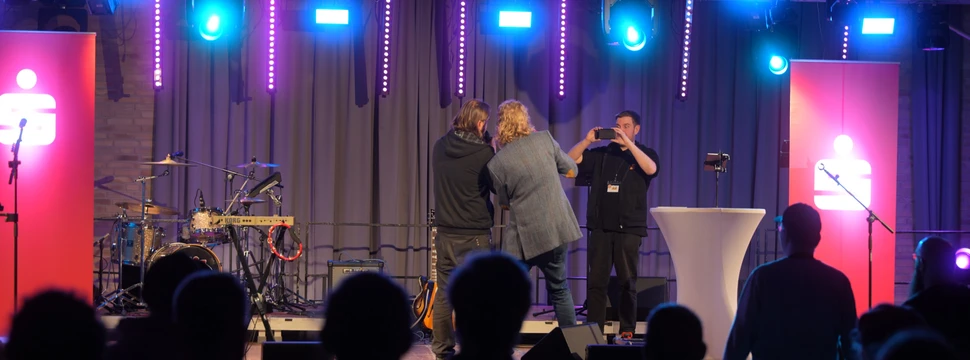 Drei Menschen auf einer Bühne vor Publikum, die gerade ein Foto machen