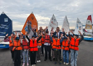 Mehrere Kinder in orangenen Rettungswesten zusammen mit mehreren erwachsenen Menschen vor der Kieler Förde bei der traditionellen Steuerradübergabe am Ende der Segelsaison
