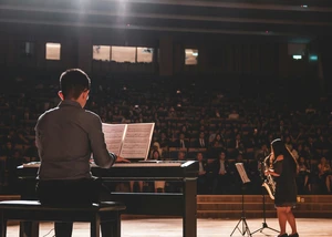 Mann sitzt mit vor einem Klavier. Im Hintergrund sieht man eine Frau am Saxophon und Publikum.