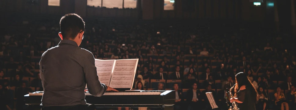 Mann sitzt mit vor einem Klavier. Im Hintergrund sieht man eine Frau am Saxophon und Publikum.